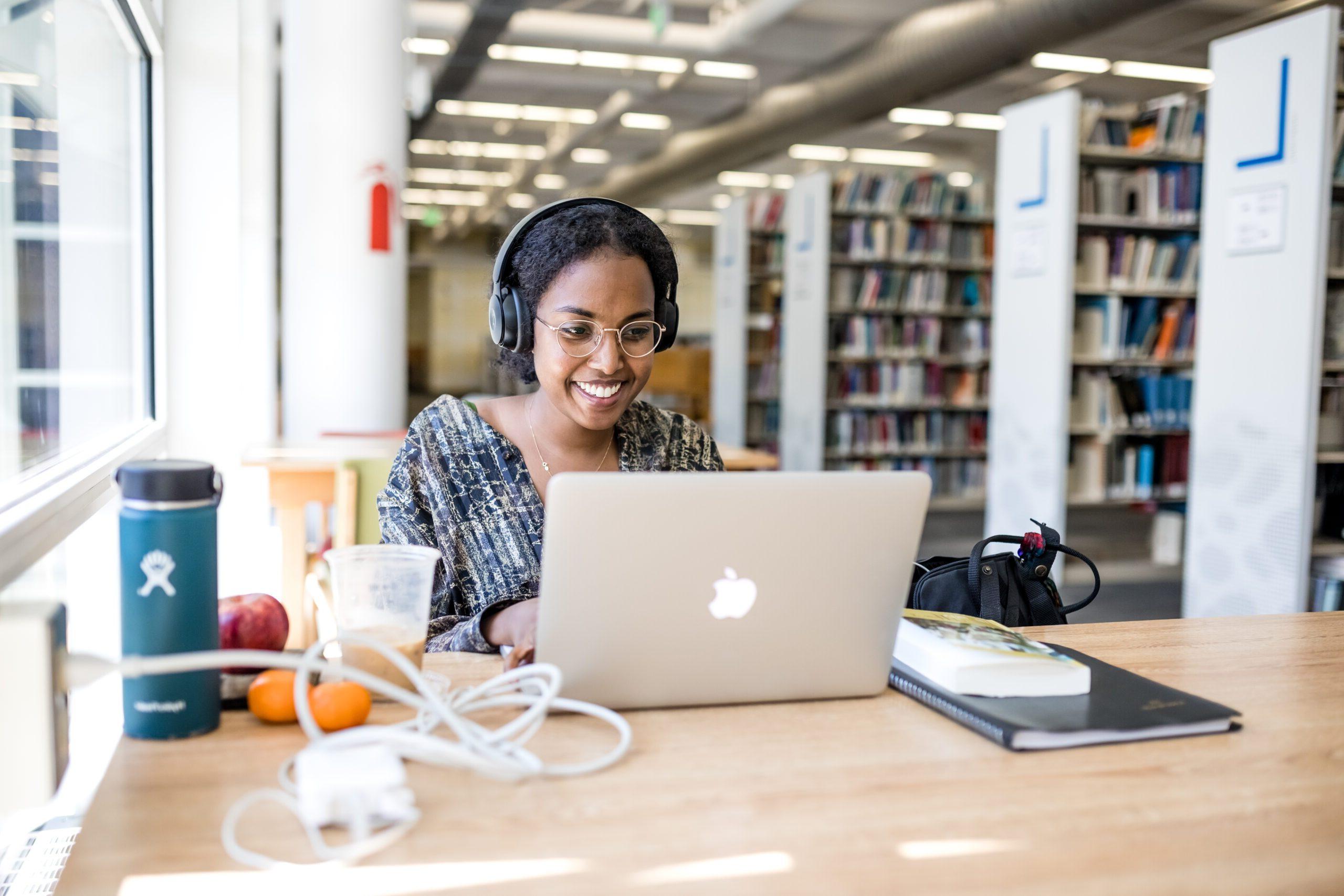 Image of a student sitting in front of a computer with headphones on in the library, smiling.